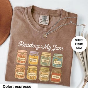 May include: Espresso-colored t-shirt with the phrase "Reading is My Jam" and illustrations of jam jars labeled with book genres. A silver necklace with a small pendant is draped across the shirt.