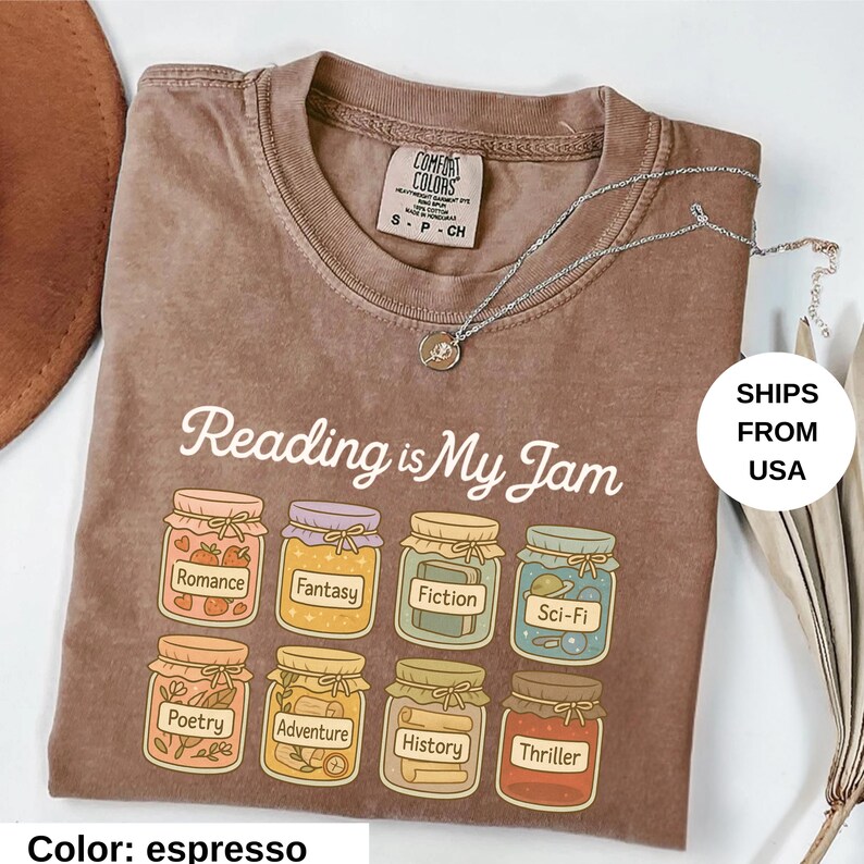May include: Espresso-colored t-shirt with the phrase "Reading is My Jam" and illustrations of jam jars labeled with book genres. A silver necklace with a small pendant is draped across the shirt.