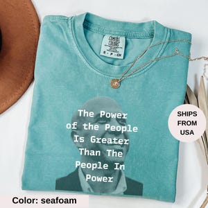 May include: Seafoam green t-shirt with the text "The Power of the People Is Greater Than The People In Power" and a portrait. A gold necklace with a pendant is on the shirt. The shirt is labeled "Comfort Colors".