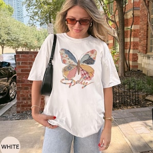 May include: A white short-sleeve t-shirt featuring a large, colorful butterfly graphic with a patchwork pattern in shades of blue, pink, and yellow. The word Dolly is written in a cursive script font underneath the butterfly design.