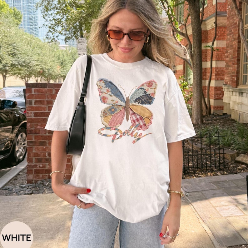May include: A white short-sleeve t-shirt featuring a large, colorful butterfly graphic with a patchwork pattern in shades of blue, pink, and yellow. The word Dolly is written in a cursive script font underneath the butterfly design.