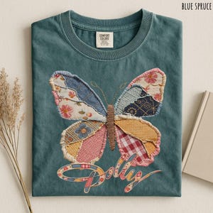 May include: A blue spruce t-shirt featuring a large butterfly graphic made of various patchwork fabric patterns including floral, gingham, and plaid prints. The butterfly has a brown embroidered body and the word Dolly is written below in script.