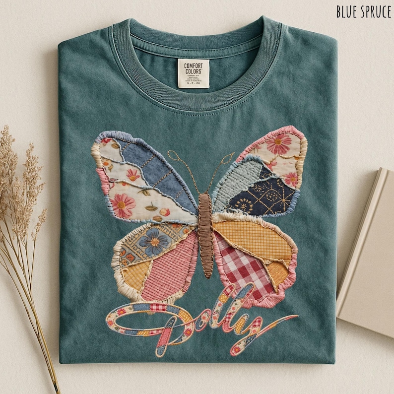 May include: A blue spruce t-shirt featuring a large butterfly graphic made of various patchwork fabric patterns including floral, gingham, and plaid prints. The butterfly has a brown embroidered body and the word Dolly is written below in script.