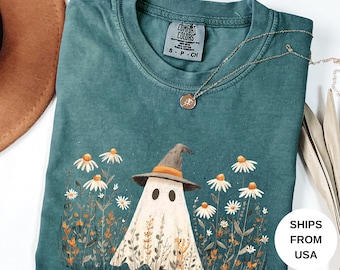 May include: Blue spruce Comfort Colors t-shirt with a ghost in a witch hat, surrounded by daisies and foliage. A gold necklace with a pendant is draped on the shirt. The shirt has the Comfort Colors logo and size information.