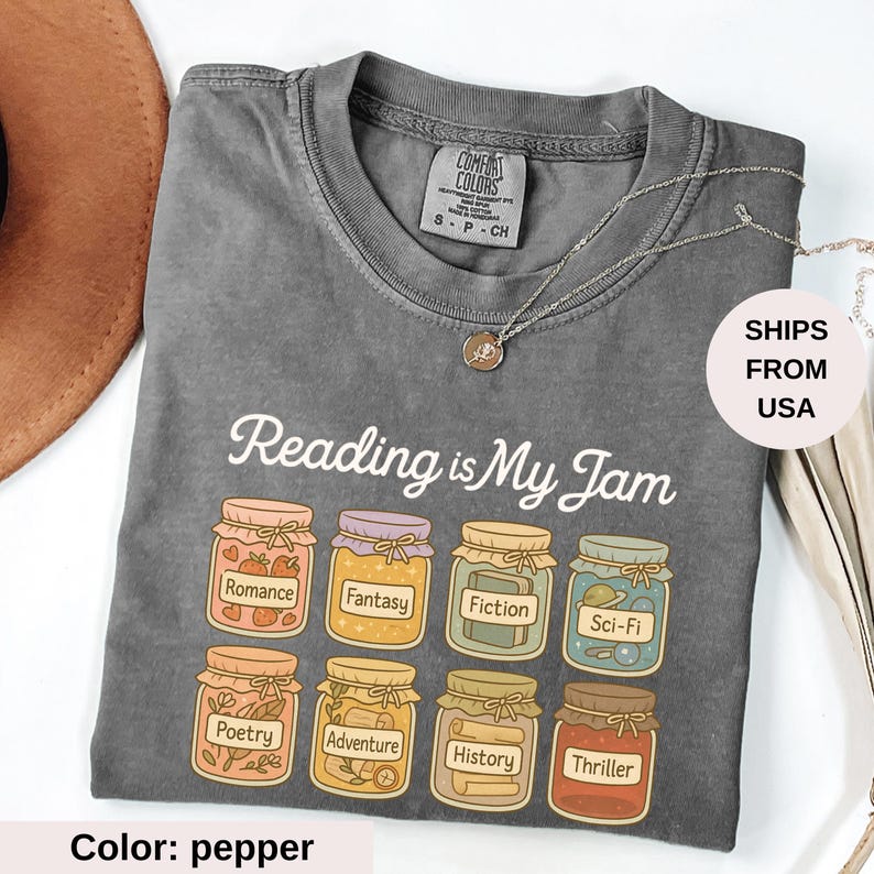 May include: Pepper-colored t-shirt with the phrase "Reading is My Jam" and illustrations of jam jars labeled with book genres. The shirt has a crew neck and short sleeves. A silver necklace is draped across the shirt.