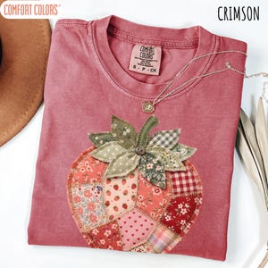 May include: Crimson Comfort Colors t-shirt featuring a patchwork strawberry design. The strawberry is crafted from diverse floral and patterned fabrics, with green leaves and a button centre. A silver necklace with a pendant is draped on the shirt.