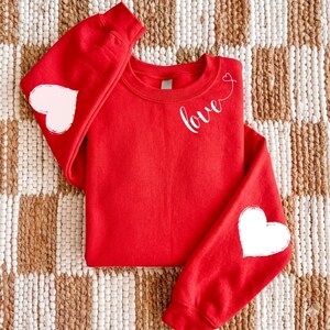 May include: A red sweatshirt with white text that says "love" and two white hearts. One heart is on each sleeve. The sweatshirt is laying on a brown and white patterned surface.
