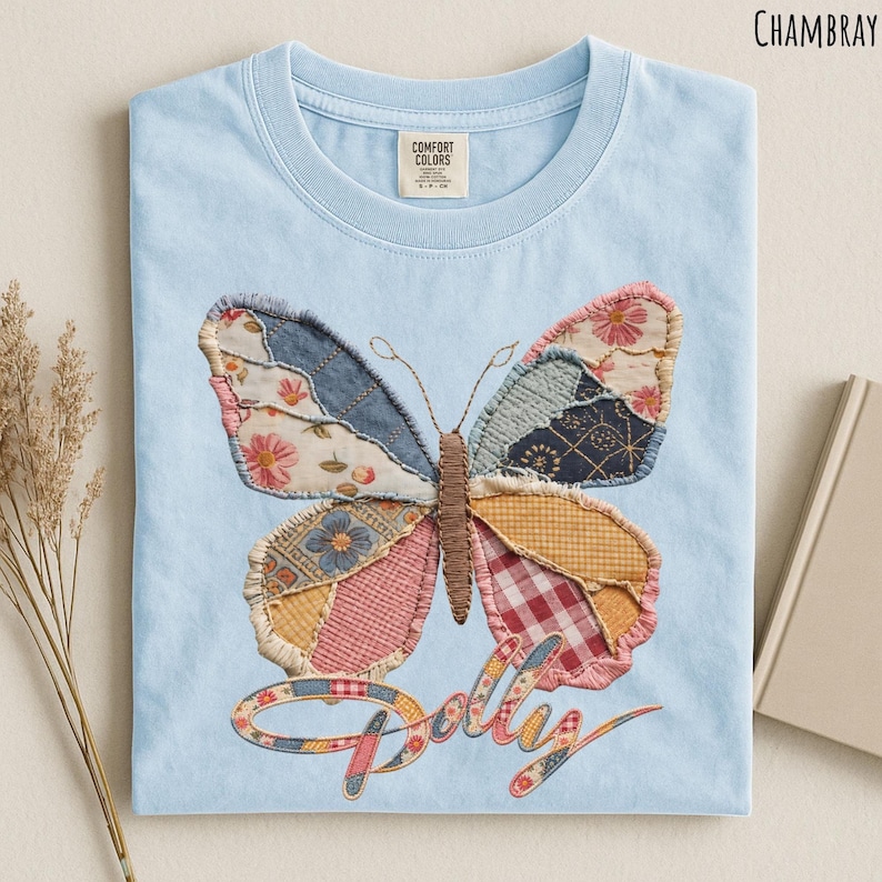 May include: A light blue t-shirt featuring a large butterfly graphic made of various patterned fabric patches in shades of pink, yellow, blue, and white. The word "Dolly" is written in a cursive script below the butterfly in a matching patchwork style.