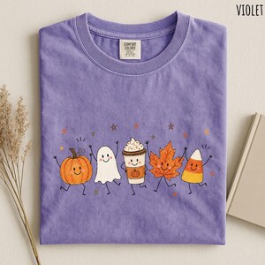 May include: A violet short-sleeved t-shirt featuring a graphic print of five smiling autumn characters: a pumpkin, a ghost, a coffee cup, a maple leaf, and a piece of candy corn, all surrounded by small stars and scattered dots.