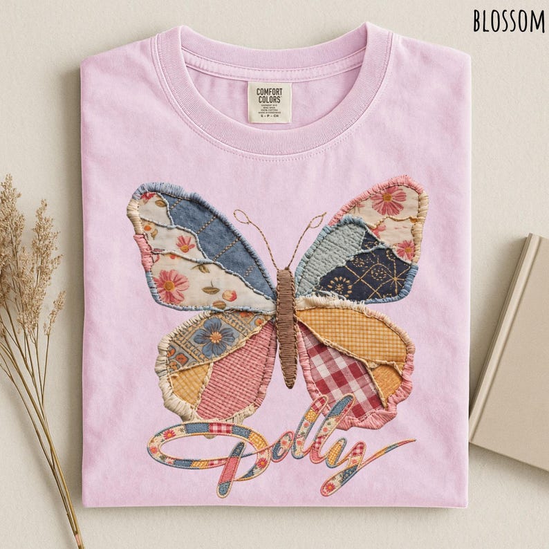 May include: A light pink t-shirt featuring a large butterfly graphic composed of various fabric patches in floral, plaid, and gingham patterns. The word "Dolly" is written in a cursive script below the butterfly, and the shirt has a crew neckline.