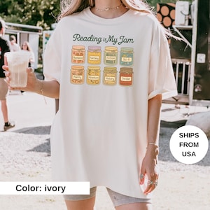 May include: Ivory-colored t-shirt with the text "Reading is My Jam" in green script. The shirt features illustrations of jam jars labeled with book genres like Romance, Fantasy, and Thriller. The shirt is a casual, comfortable style.