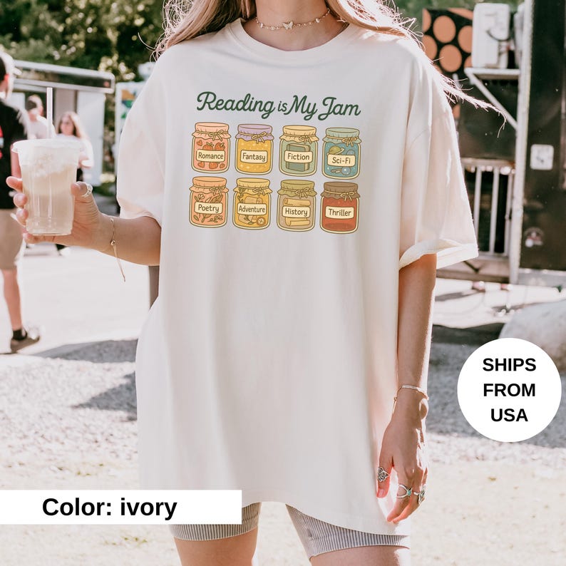 May include: Ivory-colored t-shirt with the text "Reading is My Jam" in green script. The shirt features illustrations of jam jars labeled with book genres like Romance, Fantasy, and Thriller. The shirt is a casual, comfortable style.
