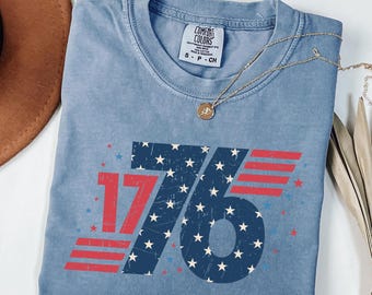 May include: A blue t-shirt with the number "1776" in a distressed design. The "76" is filled with stars and stripes, and the "17" is red. The shirt has a crew neck and short sleeves. A gold necklace is draped across the shirt.