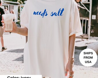 May include: Ivory-colored t-shirt with the blue text "needs salt". The shirt is a relaxed fit. The image also includes a person holding a beverage in a clear plastic cup.