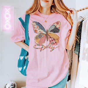 May include: A light pink short-sleeve t-shirt featuring a large butterfly graphic composed of various patchwork fabric patterns in shades of blue, red, yellow, and white. Below the butterfly, the word Dolly is printed in a cursive script font.