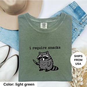 May include: Light green t-shirt with the text "i require snacks" and a cartoon raccoon holding a knife. The shirt is made of a soft material and is a comfortable fit. The shirt is displayed with a hat, jeans, and boots.