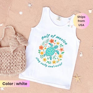 May include: White tank top with a turquoise sea turtle graphic surrounded by flowers and the text "Gulf of Mexico, stay salty and resist." A beige woven tote bag is on the left.