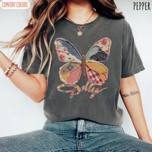 May include: A charcoal gray short-sleeve t-shirt featuring a large butterfly graphic composed of various patchwork fabric patterns in blue, pink, yellow, and red. The word Dolly is printed in a cursive script below the butterfly graphic.