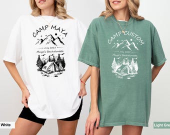 May include: White and light green t-shirts with a camping-themed graphic. The design includes mountains, a tent, trees, and the text "CAMP MAYA" or "CAMP CUSTOM", "July 2025", and "Maya's Bachelorette".