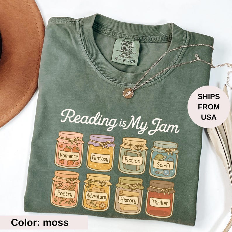 May include: Moss green t-shirt with the phrase "Reading is My Jam" and illustrations of jam jars labeled with book genres. The shirt has a crew neck and short sleeves. A silver necklace is also visible.