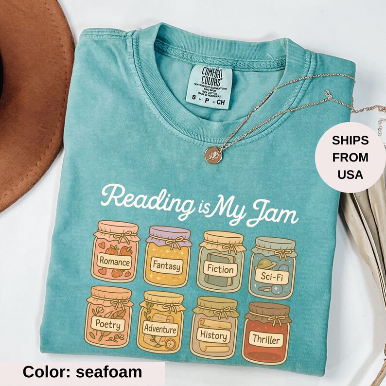 May include: Seafoam green t-shirt with the phrase "Reading is My Jam" and illustrations of jam jars labeled with book genres. The shirt has a crew neck and short sleeves. A gold necklace is draped across the shirt.