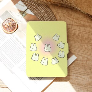 May include: A yellow tablet case featuring a pattern of white bunny faces with black outlines. The case has a soft, pastel color scheme with a pinkish center. The design is simple and cute.