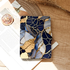 May include: A rectangular phone case with a mosaic design. The case features a pattern of navy blue, white, and gold geometric shapes. The case is lying on a wooden surface with a dried orange slice and a piece of paper.