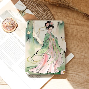 May include: A decorative notebook featuring a watercolor illustration of a woman in traditional Asian attire. The woman is wearing a flowing green and pink dress, with butterflies and flowers in the background. The notebook is rectangular.