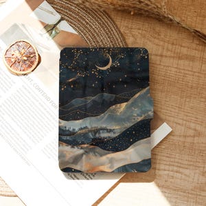 May include: A rectangular phone case featuring a celestial landscape design. The artwork depicts a dark blue night sky with a crescent moon, stars, and layered mountain ranges in shades of blue, gold, and gray.