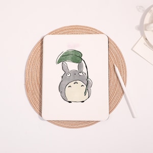 May include: A white tablet case featuring a cartoon illustration of Totoro holding a leaf umbrella. The character is gray with a white belly and black accents. The case is on a round, woven placemat with a white stylus nearby.