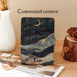 May include: A rectangular e-reader cover featuring a dark night sky with a crescent moon and gold stars above rolling hills. The design includes a flowing, layered landscape pattern in shades of blue, black, and gold. The name Kate is printed in black.