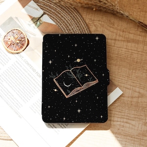 May include: A black case with a celestial design. It features an open book with a crescent moon and stars. The cover has a dark background with a gold outline of the book and illustrations. The case is rectangular and has a magnetic closure.