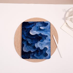 Peut inclure: Une coque de tablette avec un motif abstrait dans les tons de bleu et de blanc, rappelant des nuages ou de l'eau. L'étui a un fond bleu foncé avec des motifs tourbillonnants bleu clair et blancs. Un stylet blanc est à proximité.