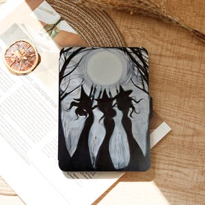 Op de afbeelding: Een zwart-grijze telefoonhoes met een griezelige illustratie van drie heksen die afsteken tegen een volle maan en een bos. De hoes heeft een matte afwerking en een beschermend ontwerp. Het thema is Halloween.