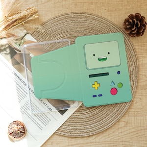 May include: A light green protective tablet case featuring a graphic of a cartoon character with a rectangular screen face, a yellow cross button, a pink circle button, a blue triangle button, and a small green circle button on the front panel.