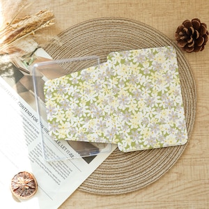 May include: A clear plastic case with a floral patterned insert. The insert features a repeating pattern of small white, yellow, and lavender flowers on a light green background. The case is on a woven placemat with a pinecone and dried citrus slice.