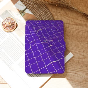 May include: A purple phone case with a white grid pattern, resembling a pool. The case is on a wooden surface with a dried orange slice and a magazine. The text "RENT, CONTEND FOR" is visible.