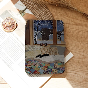 May include: A rectangular e-reader cover featuring a printed illustration of a black cat resting on a bed with a colorful patchwork quilt and floral pillows. The scene shows a snowy night view through a window with two mugs on the sill.
