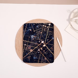 May include: A dark blue tablet case with a gold geometric design featuring lines, stars, and circles. The case is on a round, woven placemat. A white stylus is next to the case.