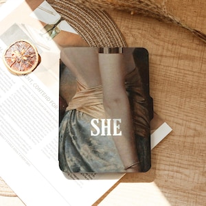 May include: A phone case featuring a classical painting of a person's back and arm, with the word "SHE" in white block letters. The person is wearing a gold-colored top and a blue skirt. A gold bracelet is visible.