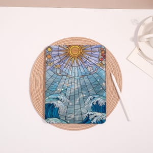 May include: A tablet case featuring a stained-glass design of a sun, clouds, and ocean waves. The artwork is predominantly blue, with yellow and white accents. A white stylus rests beside the case on a woven placemat.