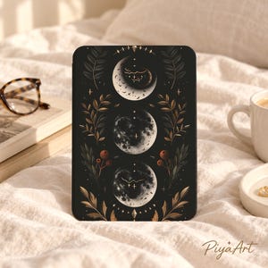 May include: A black e-reader cover featuring a vertical arrangement of three moon phases in white and gray. The design includes gold-toned botanical illustrations of leaves and berries on both sides, accented with small stars and geometric shapes.