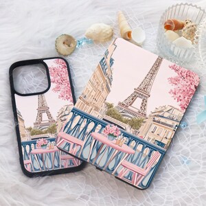May include: Two phone cases with a pink and white Eiffel Tower design. The cases feature a Parisian street scene with pink cherry blossoms and a cafe table with chairs.