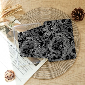 May include: A black phone case with a white topographic map design. The case is partially inside a clear plastic holder. The background includes a woven placemat, a pinecone, and a magazine with the text "DIFFERENT, CONTEND FOR THE INELE, LET IT SF."
