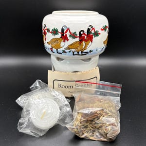 May include: A white ceramic room scenter warmer featuring a decorative band of geese wearing red bows and holly garlands. The set includes a small white tea light candle in a plastic bag and a clear plastic bag filled with dried potpourri.