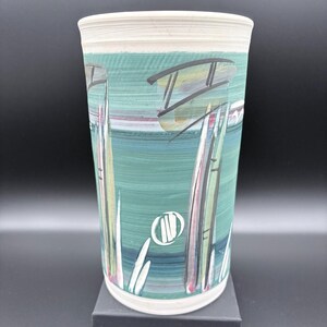 May include: A tall, cylindrical ceramic vase with a hand-painted design. The vase features a teal background with abstract shapes in black, yellow, and white. The rim is a light beige color.