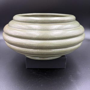 May include: A round, low-profile ceramic bowl featuring a muted sage green glaze with a subtle speckled texture. The exterior surface is defined by three distinct, horizontal, raised ridges that wrap around the circumference of the vessel.