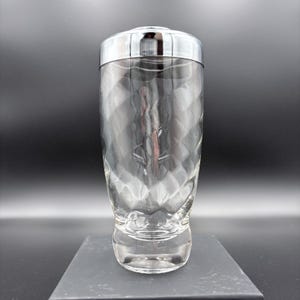 May include: A clear glass cocktail shaker with a diamond pattern and a shiny silver lid. The shaker has a wide base and a tall, cylindrical body. The lid is a simple, rounded design.