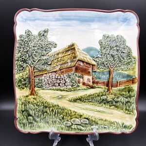 May include: Square decorative plate with a raised relief design of a rural scene. The plate features a thatched-roof barn, trees, a stone wall, and a dirt path. The plate has a scalloped edge with a brown border and is displayed on a clear stand.