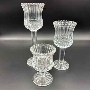 May include: Three clear glass candle holders of varying heights. Each has a tulip-shaped top with a scalloped rim and a decorative base. The tallest holder is approximately 10 inches tall, while the shortest is about 4 inches.